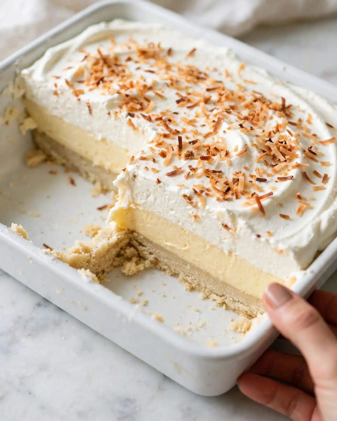 The image shows a dessert in a white rectangular dish with one square portion removed near the corner. The dessert has three visible layers: a thick, light beige base with a soft texture at the bottom, a smooth pale yellow cream layer in the middle, and a thick white whipped topping layer on top. The surface of the whipped topping is sprinkled with toasted coconut flakes that are light brown and slightly crispy. The dish is placed on a white marbled surface, and part of a woman's hand is visible holding the dish at the edge. Photo taken with an iphone --ar 4:5 --v 7